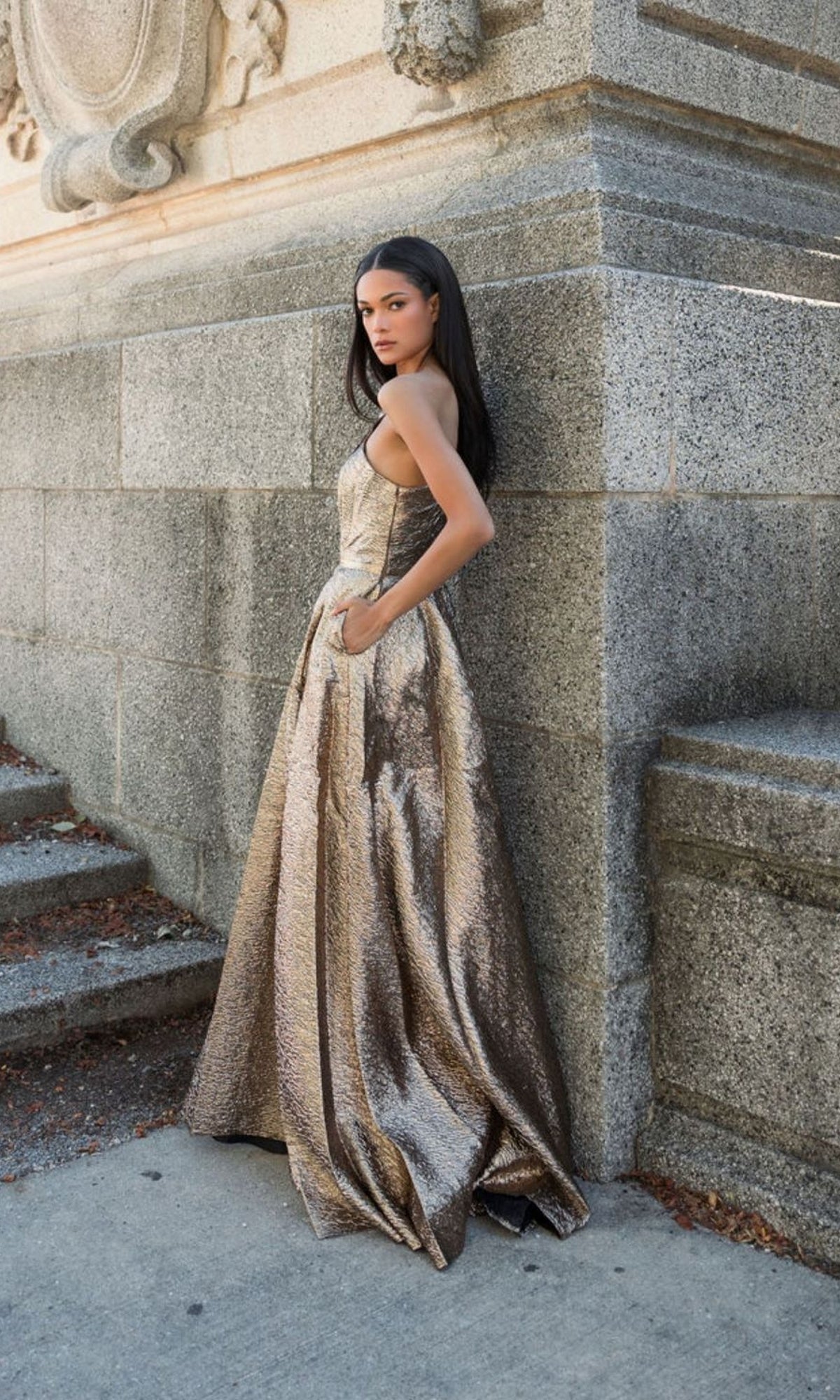 Wearing the Mac Duggal 67297 Metallic Long Prom Ball Gown, a woman poses by a stone wall on concrete steps, one hand in her pocket, gazing at the camera.